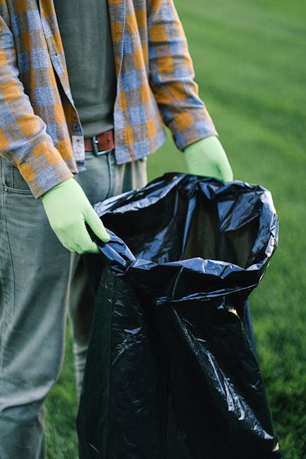 A person wearing a green long-sleeve shirt, a plaid jacket with yellow and blue tones, and light grey trousers with a brown belt, is standing outdoors on a grassy area. They are holding open a large b