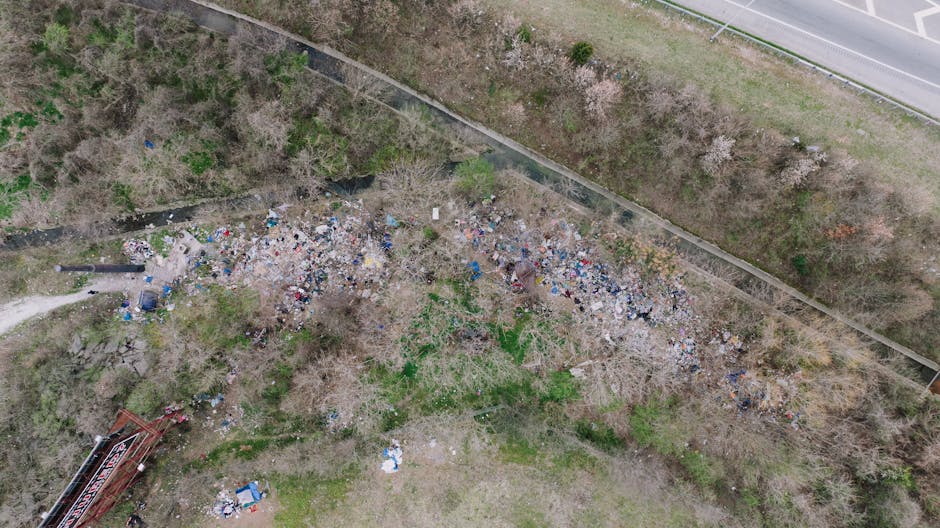 A large collection of mixed domestic waste is piled on a paved area in front of a recycling station, comprising various black, red, and gray bins, some overflowing with black and white plastic bags, c